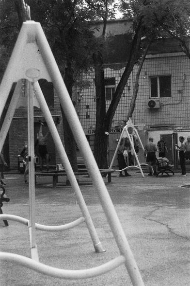 ORWO P400 (FF)

English Alt Text: A black-and-white image of a playground with a large triangular metal swing frame in the foreground, though no swings are attached. In the background, another similar frame is surrounded by people—adults and children—engaging in conversation or play. Benches and trees are scattered throughout. A tiled building with windows and air conditioning units stands behind the scene. The image captures a quiet moment of urban recreation.

中文替代文字：
这是一张黑白照片，拍摄于一个游乐场。前景是一座大…