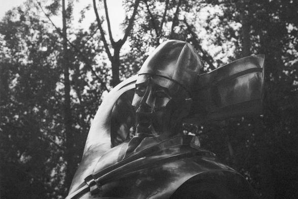 Ilford HP5 Plus 400 (FF)

English Alt Text:
A black and white photo of a metallic sculpture resembling a stylized human figure, possibly a knight or warrior. The sculpture features angular, abstract shapes. Its surface is smooth and reflective, catching light in dramatic ways. The background consists of tall, shadowy trees, adding depth and contrast to the scene. The overall tone is solemn and artistic, emphasizing the craftsmanship and expressive form of the sculpture.

中文替代文字：
这是一张黑白照片，展示了一个金…