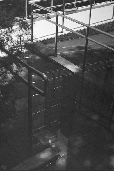 Ilford HP5 Plus 400 (FF)

English Alt Text:
A monochrome photograph showing metal rails by the side of  a dark, reflective puddle. Railings surround the area, mounted on a tiled or concrete surface. The water reflects the railings, and nearby trees, creating layered shadows and geometric patterns. The composition emphasizes contrast between light and dark, with a quiet, contemplative mood.

中文替代文字：
这是一张黑白照片，画面中金属栏杆在深色、反光的水洼的旁边。水洼周围有金属栏杆，安装在瓷砖或混凝土表面上。水面映出栏杆和周围树木的倒影，形成层次分明的阴影和几何图案。画面强调明暗对比，营造出宁静、…