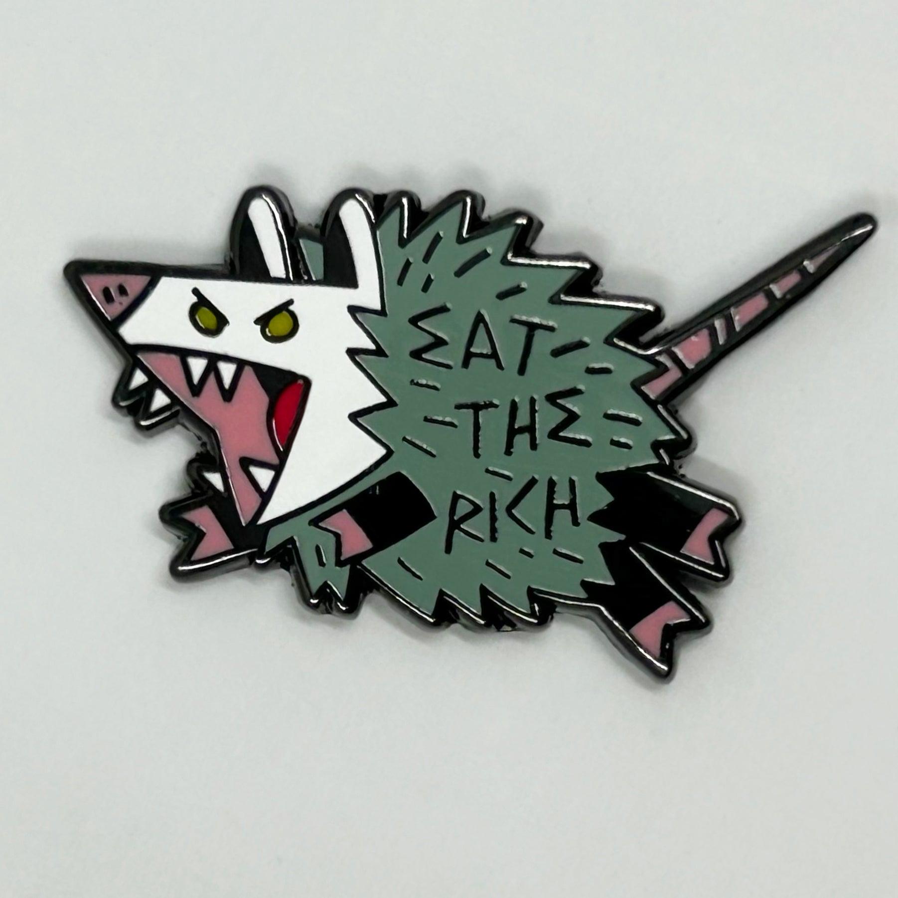 A pin of a possum that says Eat The Rich.