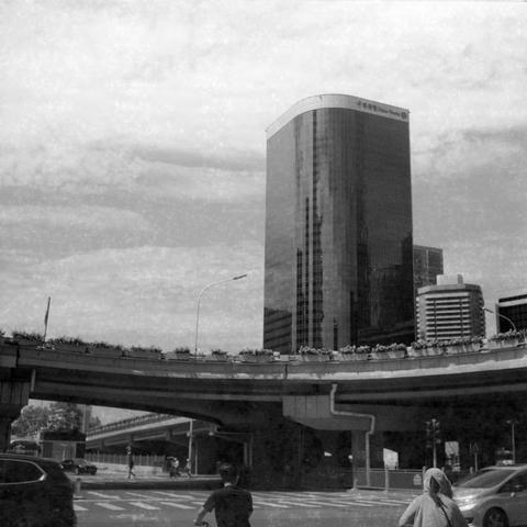 Ilford HP5 Plus 400 (6x6)

English Alt Text:
A black-and-white urban scene dominated by a tall skyscraper with reflective glass windows and a curved facade. Chinese characters are displayed at the top of the building. In the foreground, an elevated highway runs across the frame, with greenery along its edge. Below, a road with painted traffic markings is visible. Two people, one on a bicycle and one walking, move toward the underpass. The photo captures the scale of modern architecture against …