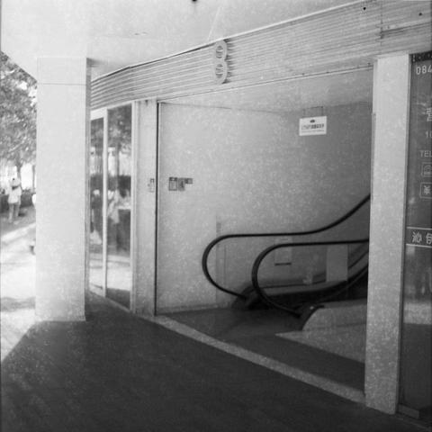 Ilford HP5 Plus 400 (6x6)

English Alt Text:
A monochrome image of a modern building entrance with a glass facade. Inside, an escalator descends from the upper floor to the ground level. Above the entrance and on the interior wall, signage with Japanese text is visible. Outside, people walk along the sidewalk shaded by trees. The grainy texture suggests the photo was taken with film or is an older print. The composition captures the contrast between movement—people and escalator—and the stillne…