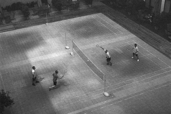 Ilford HP5 Plus 400 (FF)

English Alt Text:
A black-and-white overhead view of four people playing badminton on a marked court. The net divides the space, and each player holds a racket, actively engaged in the game. One player is captured mid-swing, striking the shuttlecock. Tables and chairs in the background suggest a courtyard or recreational area. The image conveys energy, movement, and social interaction within a structured play environment.

中文替代文字：
一张黑白照片，从上方俯拍四个人在羽毛球场上比赛。球网将场地分为两半，每位选手…