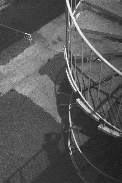 Ilford HP5 Plus 400 (FF)

English Alt Text:
A black-and-white photograph of a spiral metal staircase viewed from above. The staircase curves downward in a clockwise direction, casting a shadow on the concrete ground below. Patches of grass break up the pavement, and a railing is visible in the corner, suggesting another walkway. The interplay of curves, shadows, and textures creates a visually dynamic and geometric composition.

中文替代文字：
一张黑白照片，从上方俯视一座金属螺旋楼梯。楼梯顺时针向下延伸，在混凝土地面上投下弯曲的阴影。地面上点缀着草地，左上角…