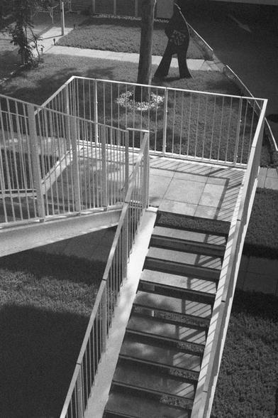 Ilford HP5 Plus 400 (FF)

English Alt Text:
A black-and-white photograph of a metal outdoor staircase leading up to a landing. The stairs and railings create sharp geometric shadows on the ground and wall, forming a striking pattern of light and dark. Grass and a sidewalk surround the structure, while a tree stands nearby. In the background, a sculpture shaped like a human figure with outstretched arms adds an artistic element. The composition emphasizes contrast, geometry, and the play of sunl…