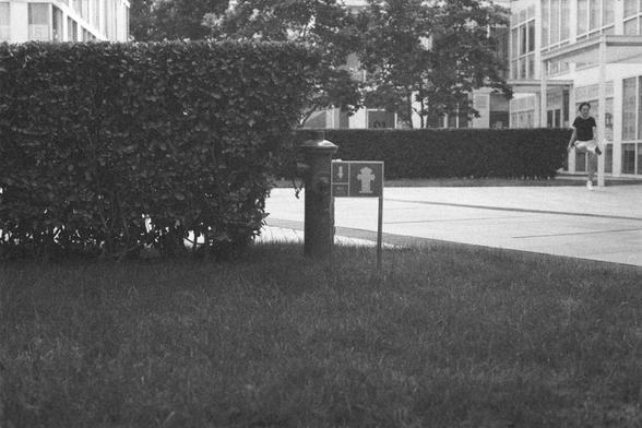 Ilford HP5 Plus 400 (FF)

English Alt Text:
A black-and-white photograph of an outdoor urban scene. In the foreground, a neatly trimmed hedge borders a grassy area. A small sign with a fire hydrant symbol and a downward arrow marks the hydrant’s location beside the grass. The hydrant itself is visible near the sign. Behind this, modern buildings with large windows rise, and a paved walkway runs alongside them. A person is faintly visible near the buildings, adding a sense of scale. The photo em…