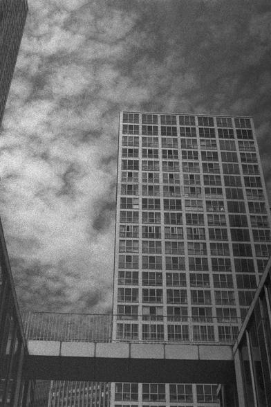 Ilford HP5 Plus 400 (FF)

English Alt Text:
A black-and-white cityscape featuring tall modern buildings. The central high-rise has a grid of square windows, creating a repetitive geometric pattern. In the foreground, a pedestrian bridge connects two buildings, adding depth and perspective. Above, textured clouds fill the sky, enhancing the dramatic mood. The low-angle viewpoint emphasizes the towering scale of the architecture.

中文替代文字：
一张黑白城市景观照片，展示高耸的现代建筑。中央的高楼布满方形窗户，形成规则的几何图案。前景是一座连接两栋建筑的人行天…