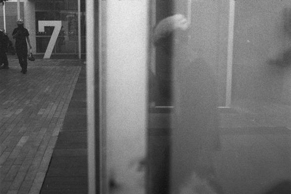 Ilford HP5 Plus 400 (FF)

English Alt Text:
A grainy black-and-white urban photograph, possibly taken through glass. On the left, a person wearing a helmet and carrying a bag walks along a wooden or tiled pathway. Behind them, a large number “7” is displayed on a glass wall or door, suggesting a building entrance or floor marker. The right side of the image is blurred with reflections, showing indistinct shapes and possibly another figure. The photo has a vintage, film-like texture, emphasizing…