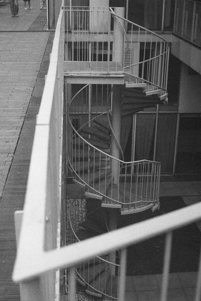 Ilford HP5 Plus 400 (FF)

English Alt Text:
A black-and-white photograph of a spiral metal staircase enclosed between two building walls. The staircase curves downward in a tight coil, with railings tracing its circular form. The perspective is from above, looking down into the narrow vertical shaft. The repeating lines of steps and railings create a hypnotic geometric pattern. The photo emphasizes symmetry, depth, and the interplay of light and shadow on the metal surfaces.

中文替代文字：
一张黑白照片，画面中…