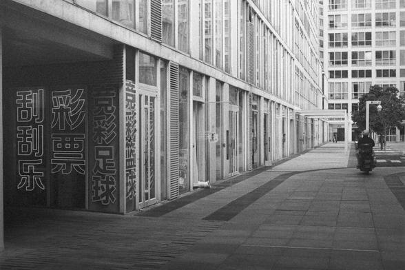 Ilford HP5 Plus 400 (FF)

English Alt Text:
A modern city street lined with tall glass-and-metal buildings. Large windows with vertical blinds reflect light, creating a sleek, geometric look. On the left, several brightly colored vertical signs in Chinese characters stand out against the grayscale tones. One sign reads “lottery,” another “football,” suggesting shops or betting outlets. A cyclist or motorbike rider moves away from the camera on the right side of the tiled street. Trees peek thro…