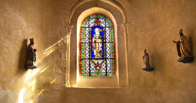 A stained glass window with light streaming through it. Small figures of saints are on shelves on the wall. 