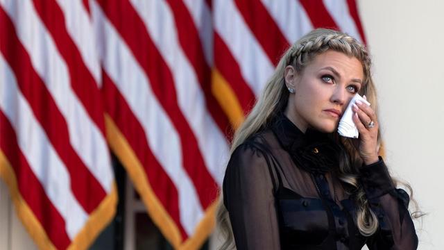 A blond woman in a sheer top holding a folded cloth near her eyes in front of three American flags.