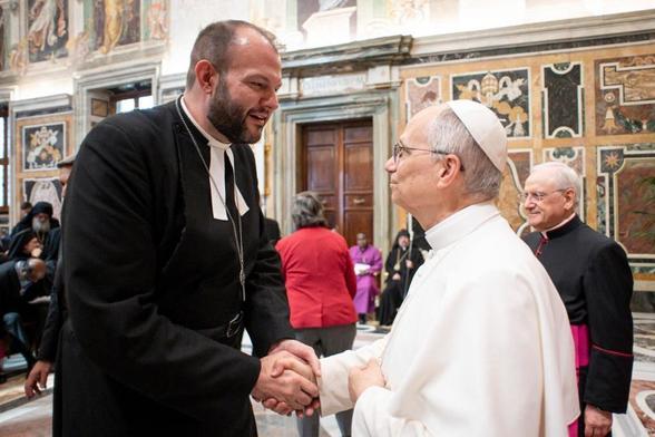 A tall man in priestly garb shaking hands with the pope.