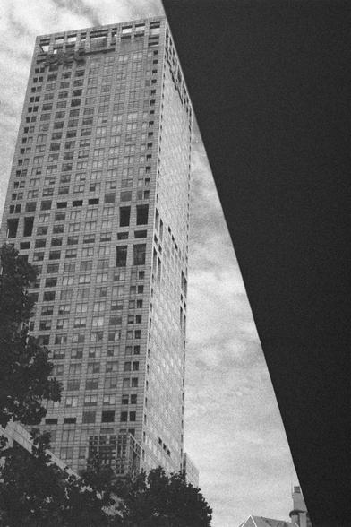 Ilford HP5 Plus 400 (FF)

English Alt Text:
A tall high-rise building with many windows, viewed from below to emphasize vertical lines. A large dark structure, possibly an overpass or another building, cuts diagonally across the right side, creating a dramatic framing effect. At the bottom, trees and smaller buildings add depth to the urban setting. The sky is partly cloudy, and the overall tone is moody, with strong contrasts between light and shadow.

中文替代文字：
一栋高层建筑从低角度拍摄，窗户密集，垂直线条突出。画面右侧有一处巨…