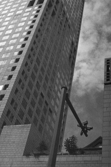 Ilford HP5 Plus 400 (FF)

English Alt Text:
A tall modern skyscraper photographed from a low angle, emphasizing its height and grid-like windows. In the foreground, a security camera mounted on a pole points upward, suggesting surveillance of the building and surrounding area. The sky is partly cloudy, adding texture to the background. At the base, smaller structures and vegetation are visible. The perspective highlights both the scale of the building and the presence of urban monitoring.

中文替代…