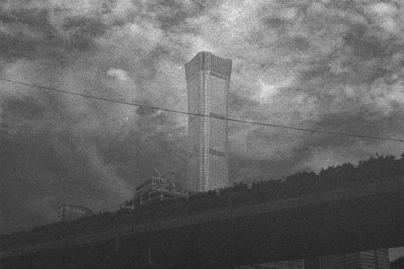Ilford HP5 Plus 400 (FF)

English Alt Text:
A uniquely shaped skyscraper dominates the skyline, with an hourglass-like design that is wider at the top and bottom but narrow in the middle. The photo is in black and white with a grainy texture, giving it a vintage or film noir feel. In the foreground, an elevated highway with vegetation runs across the frame, while smaller buildings and construction appear near the base. A power line cuts diagonally across the image. The dramatic sky is filled wi…