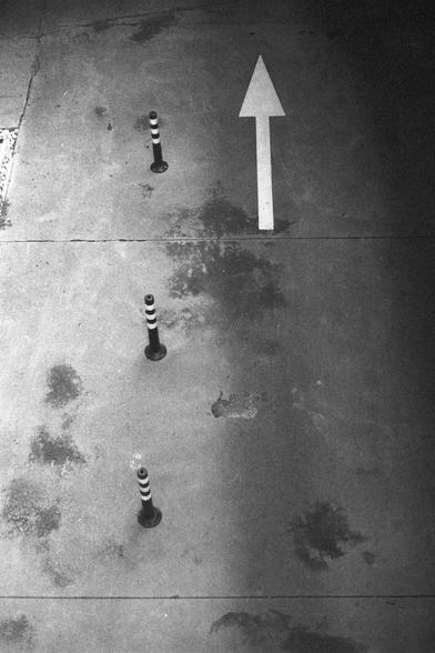 Ilford HP5 Plus 400 (FF)

English Alt Text:
A concrete pavement with a large white arrow painted on the ground, pointing forward to indicate direction. On the left side of the arrow, four short black-and-white striped bollards stand in a row, acting as barriers or guides. The pavement surface shows stains and marks from frequent use. The right side of the image is darker due to shadow, while the left side is brighter, creating contrast. The scene suggests an urban walkway or vehicle path with c…