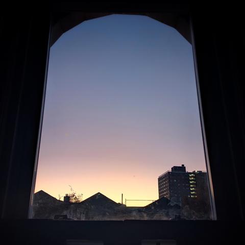 Looking through an arched window twenty-three minutes before sunrise the cloudless sky fades from pale yellow at the horizon to dusty blue up above. Pointed roofs of Harlem brownstones are silhouetted across the street, and a taller apartment building can be seen in the distance. 