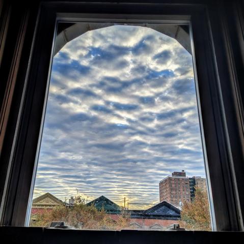 Looking through an arched window forry-six minutes after sunrise the sky is filled with striated clouds: a mackerel sky. The sun is obscured, but creating a yellow tinge at the horizon. Pointed roofs of Harlem brownstones are across the street, and a taller apartment building can be seen in the distance. The tops of two trees on the bottom and right are turning gold.