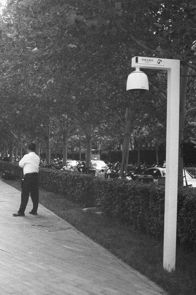 Ilford HP5 Plus 400 (FF)

English Alt Text:
A black-and-white photo of a city sidewalk. A man stands with his back to the camera, facing the street. Trees and a hedge line the walkway, separating it from the road. Several motorcycles and cars are parked or moving along the street. On the right, a tall white pole supports a dome-shaped surveillance camera. Above the camera, a sign with Chinese characters reads “Video Surveillance.” The atmosphere feels urban, orderly, and slightly watchful, emph…