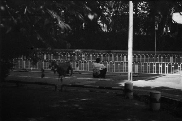ERA 100 (FF)

English Alt Text:
A black-and-white photograph shows two people on a quiet sidewalk beside a road. One person sits on the ground with knees bent and arms resting on their legs, facing away from the camera. The other person crouches nearby, leaning slightly forward. A white metal fence separates the sidewalk from a tree-lined area in the background. Shadows from trees and structures fall across the pavement, creating strong contrasts of light and dark. The mood feels contemplative,…
