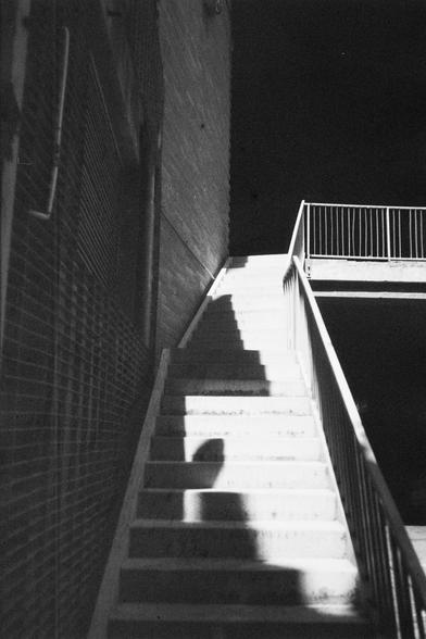 ERA 100 (FF)

English Alt Text:
A dramatic black-and-white photo of a staircase at night. A strong light illuminates the steps, casting a clear shadow of a person standing at the base. The shadow stretches upward across the lit stairs, creating a mysterious silhouette. The staircase leads to a landing with a metal railing, and the surrounding walls are brick. The interplay of light and shadow emphasizes architectural lines and evokes a moody, cinematic atmosphere, suggesting presence without re…