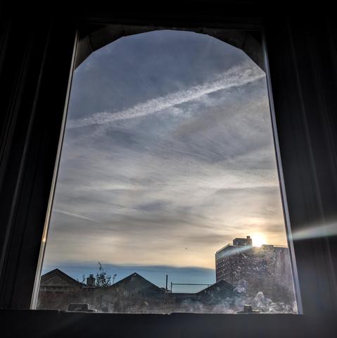 Looking through an arched window ninety minutes after sunrise the sky is filled with horizontal slashes of grey and white clouds. The pale yellow sun is just above the houses on the right, creating a diagonal white line of lens flare. There is a band of indigo at the horizon. Pointed roofs of Harlem brownstones are silhouetted across the street, and a taller apartment building can be seen in the distance. The window is grimy on the bottom.