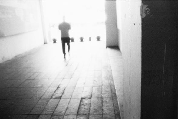 ERA 100 (FF)

English Alt Text:
A black-and-white photograph of a tiled corridor. A single person walks away from the camera toward a bright opening at the far end, appearing as a dark silhouette against the strong backlight. The figure is slightly blurred, suggesting motion. The corridor walls are dim, with faint text in a non-Latin script visible on the right side. The contrast between the dark passage and the glowing exit creates a dramatic sense of depth, mystery, and anticipation.

中文替代文字：…