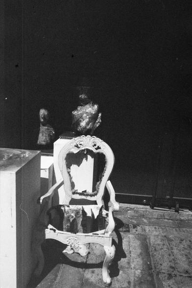 ERA 100 (FF)

English Alt Text:
A black-and-white photograph showing a broken ornate chair in the foreground. The chair’s decorative frame remains, but its seat and parts of the backrest are missing, exposing its inner structure. Behind it, two sculpted busts rest on a pedestal, partially hidden in shadow. The background is dark, contrasting with the illuminated chair. The scene evokes themes of decay, time, and art, with the fragile chair juxtaposed against classical sculptures.

中文替代文字：
一张黑白照…