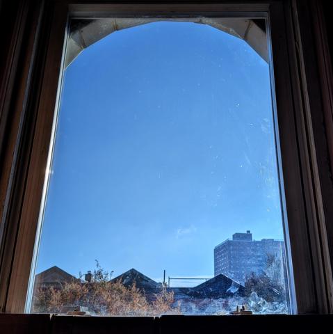 Looking through an arched window three and a half hours after sunrise the bright blue sky is marked with a few subtle wisps of white cloud. The sun is out of frame above the buildings on the right. Pointed roofs of Harlem brownstones are silhouetted across the street, and a taller apartment building can be seen in the distance. The tops of two brown trees are on the bottom and right. The window is grimy on the bottom.
