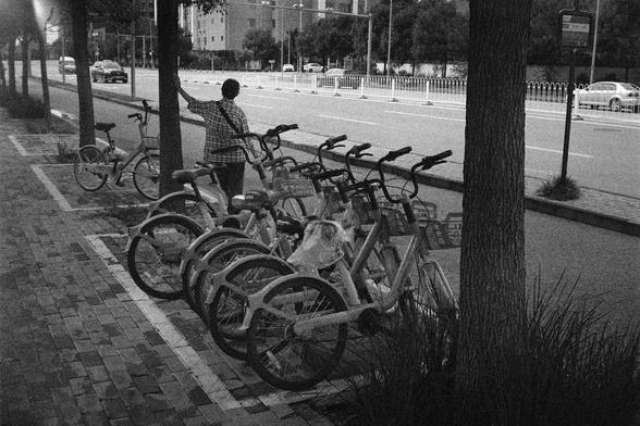 Shanghai GP3 400 PAN (FF)

English Alt Text:
A monochrome photo of a sidewalk lined with uniform bicycles, likely part of a bike-sharing system. Each bicycle has a QR code plate attached. A person stands beside one bicycle, facing the street, as if preparing to ride. Across the multi-lane road, cars and buildings are visible. Trees line the sidewalk, and the pavement is made of rectangular bricks. The grainy black-and-white style gives the image a vintage or surveillance-like quality, emphasizi…