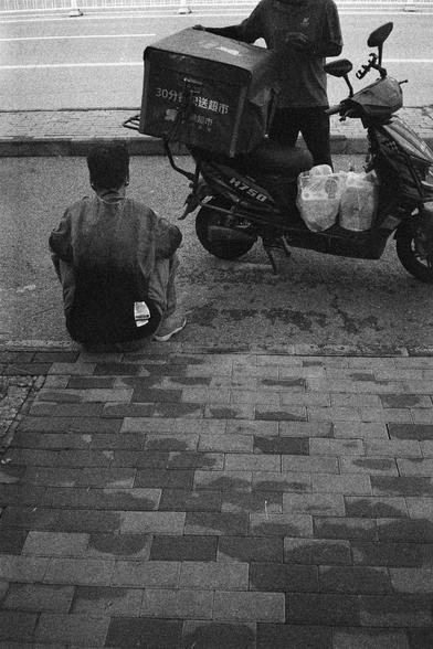 Shanghai GP3 400 PAN (FF)

English Alt Text:
A black-and-white photograph of a city sidewalk. In the foreground, a person sits on the pavement with their back to the camera, appearing to rest or wait. Ahead, another person stands beside a motor scooter equipped with a large delivery box. The box has Chinese text that translates to “30-minute supermarket delivery.” Several plastic bags hang from the scooter, likely filled with groceries. The curb and road are visible in the background, suggestin…