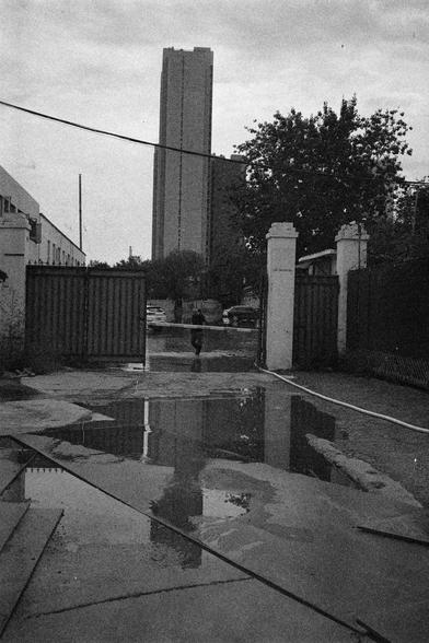 Shanghai GP3 400 PAN (FF)

English Alt Text:
A gritty black-and-white urban scene. A wet pavement dominates the foreground, with large puddles reflecting nearby structures. An open gate with two white pillars leads into a parking area where several cars are parked. A person walks away from the camera toward the gate. In the background, a tall building rises above smaller structures, framed by trees and overhead wires. The grainy texture and reflections create a vintage, documentary atmosphere, …