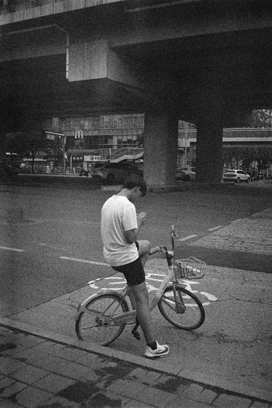 English Alt Text:
A grainy black-and-white photo of a person sitting on a shared bicycle under a city overpass. The person wears a white t-shirt, dark shorts, and sneakers, and is looking at a mobile phone. The bike has a front basket. Behind them, cars drive along the road, and a McDonald’s restaurant is visible among other buildings. The scene captures a candid moment of modern urban life, blending technology, transportation, and solitude.

中文替代文字：
这是一张黑白颗粒感照片，一位骑共享单车的人坐在城市高架桥下。他穿着白色T恤、深色短裤和运…