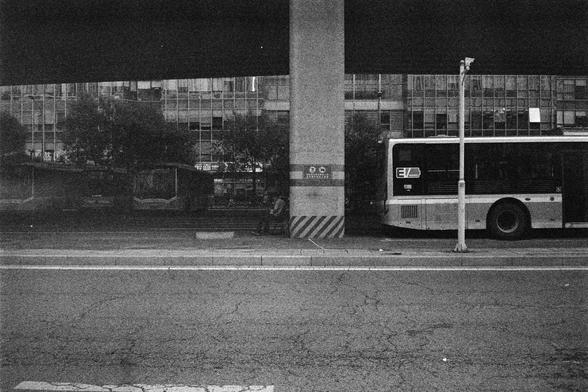 Shanghai GP3 400 PAN (FF)

English Alt Text:
A grayscale image of a city bus stop located beneath a concrete overpass. A large pillar supports the structure above, dividing the scene. On the right, a bus is parked near the curb; another bus is visible in the distance. A person sits alone on a bench near the pillar, possibly waiting. Behind them, a modern glass building and trees line the sidewalk. The cracked pavement and faded crosswalk add texture. The monochrome palette evokes a quiet, conte…
