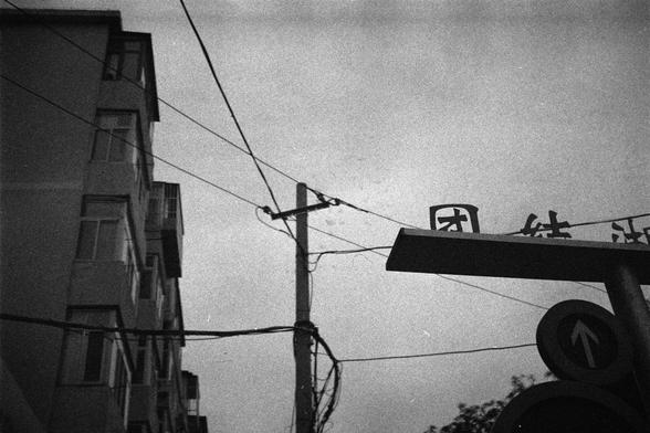 Shanghai GP3 400 PAN (FF)

English Alt Text:
A black-and-white photo of an urban street scene dominated by a tall utility pole in the center. Numerous electrical wires stretch outward in various directions, forming a web-like pattern against an overcast sky. To the left, a multi-story building with rectangular windows stands quietly. On the right, a partial sign with Chinese characters and a circular traffic sign featuring an upward-pointing arrow suggest a one-way street. The mood is somber an…