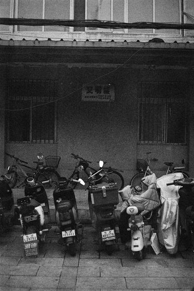 Shanghai GP3 400 PAN (FF)

English Alt Text: A black-and-white street photo showing a cluttered scene of parked motor scooters and bicycles in front of a building. Some scooters have license plates, others are covered with protective sheets. The building behind has barred windows and a sign with Chinese characters above the entrance. The image captures the density and texture of urban life, with overlapping shapes and surfaces creating a documentary-style composition.

中文替代文字：
这是一张黑白街头照片，画面前景是密…