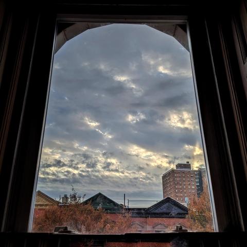 Looking through an arched window an hour after sunrise the sky is filled with chunky gray clouds, and patches of yellow light are breaking through in a few places. Pointed roofs of Harlem brownstones with red brickwork are across the street, and a taller apartment building can be seen in the distance. The tops of two trees are on the bottom and right, turning red and gold for autumn.