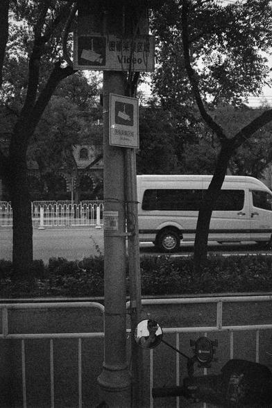 Shanghai GP3 400 PAN (FF)

English Alt Text: A monochrome street scene centers on a utility pole with two signs showing a surveillance camera icon and the word “Video” in English and Chinese (图像采集区). These signs indicate the area is under video surveillance. A round mirror is attached to the pole. In the background, a white van is visible near trees and a building with arched windows. In the foreground, part of a scooter or bike is seen, with a device labeled “innotek” on the handlebar.

中文替代文字…