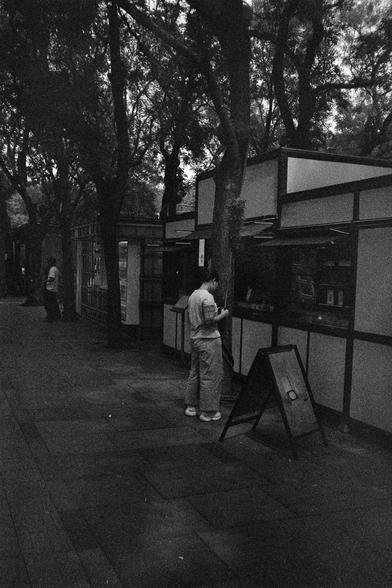 Shanghai GP3 400 PAN (FF)

English Alt Text: A black-and-white photo shows a quiet urban street shaded by tall trees. A person stands in front of a small kiosk or shop with a traditional design, featuring rectangular panels and a counter window. The person appears to be reading a menu or placing an order. A freestanding signboard is positioned in front of the shop. Another person walks further down the tiled walkway. The scene captures a peaceful moment of daily life, with grainy texture sugges…