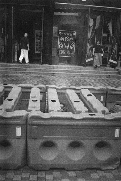Shanghai GP3 400 PAN (FF)

English Alt Text: A grainy black-and-white photo shows a storefront with a large sign reading “NOFAKES” and Chinese text “奢侈品 回收寄卖”, meaning “Luxury goods recycling and consignment.” The sign includes illustrations of a handbag, necklace, and watch. In front of the store, rows of large plastic barriers are arranged, possibly for crowd control or construction. People are seen entering and exiting the store, suggesting active business.

中文替代文字：
这是一张颗粒感强烈的黑白照片，展示了一家店铺的门面…