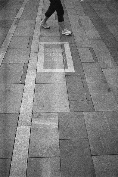 Shanghai GP3 400 PAN (FF)

English Alt Text: A black-and-white image shows a person walking on a tiled pavement. The person wears athletic shoes and cropped pants. The pavement consists of rectangular tiles in various shades of gray, arranged in a linear pattern. Near the center, a rectangular embedded marker with faded Chinese text is visible. The characters are partially legible, possibly reading “…安”, “…生”, “…全”, suggesting a message about safety or public welfare.

中文替代文字：
这是一张黑白照片，展示了一个人走在…