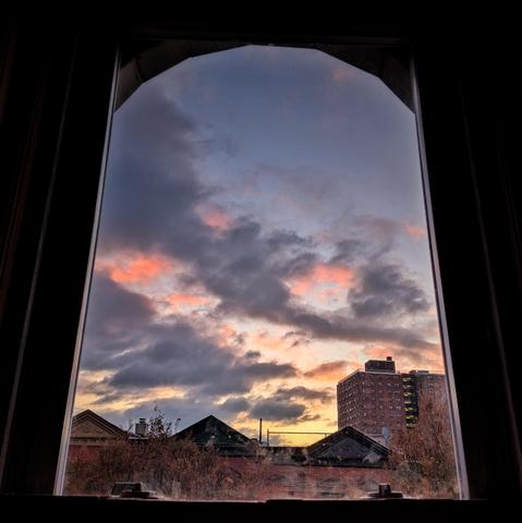 Looking through an arched window one minute after sunrise the dusty blue sky is splashed with gray clouds underlit with bright pink. A pool of yellow sunlight is at the horizon. Pointed roofs of Harlem brownstones are across the street, and a taller apartment building can be seen in the distance. The tops of two autumnal trees are on the bottom and right.