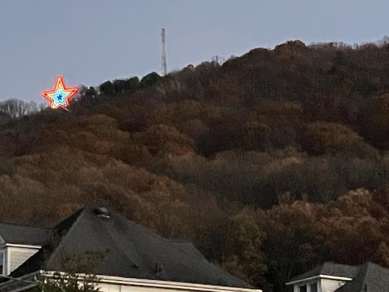 A brightly lit star, featuring a blue center and outlined in red and orange, is positioned on a mountain against a backdrop of trees with autumn foliage. In the foreground, rooftops of nearby houses are visible.