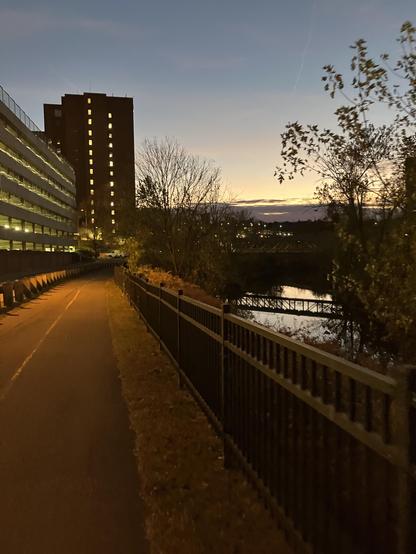 A pathway beside a river at dusk, with buildings and trees on either side. The sky displays a gradient of colors as the sun sets, and a metal fence runs along the path.