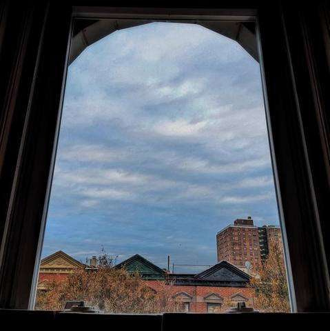 Looking through an arched window twenty-four minutes after sunrise the powder-blue sky is full of undulating, diffuse white clouds. Pointed roofs of Harlem brownstones with red brickwork are across the street, and a taller apartment buildifng can be seen in the distance. Two trees with brown leaves are on the bottom and right.