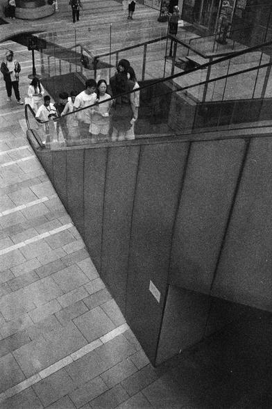 Shanghai GP3 400 PAN (FF)

English Alt Text: A group of people ride an upward-moving escalator enclosed by glass panels. The architecture features tiled floors and angular surfaces. On the upper level, more people walk in the background. The image captures a moment of daily urban life with geometric design and human movement.

中文替代文本： 这是一张黑白照片。几位乘客正乘坐向上的自动扶梯，扶梯被玻璃护栏包围。建筑设计呈现出瓷砖地面和锐角结构。上层背景中还有其他人在行走。画面展现了城市日常生活的一瞬，几何建筑与人群动态交织。