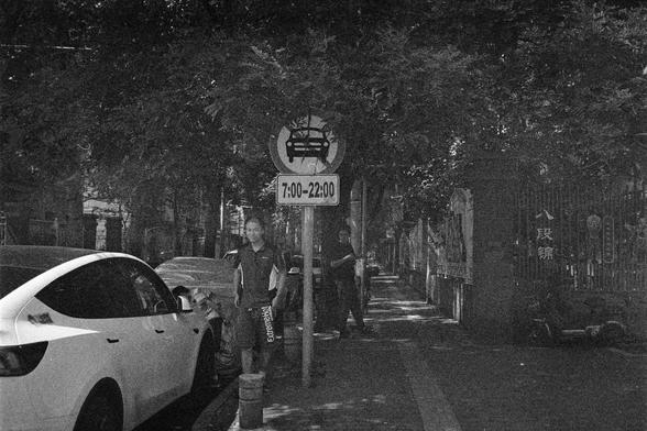 Shanghai GP3 400 PAN (FF)

English Alt Text: A black-and-white photo of a tree-lined urban street. On the sidewalk, several pedestrians walk past parked cars. A traffic sign is prominently displayed: a red circle with a diagonal line over a car symbol, indicating no vehicle entry. Below it, the restricted hours are listed as “7:00 - 22:00.” In the background, buildings and a fence are visible. The fence bears Chinese characters “八段锦,” referring to a traditional qigong exercise. The scene blends…