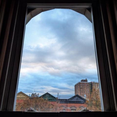 Looking through an arched window nineteen minutes after sunrise the candy blue sky is filled with rolling, wintery white clouds. Pointed roofs of Harlem brownstones with red brickwork are across the street, and a taller apartment building can be seen in the distance. The tops of two trees are on the bottom and right, turning red and brown for autumn.
