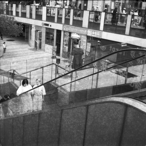 Ilford HP5 Plus 400 (6x6)

English Alt Text: A black-and-white photo of a bustling multi-level structure. The lower level features a glass-paneled escalator and stairway, with people ascending and descending. The upper level has storefronts with Chinese signage and individuals walking along the corridor. Reflections on glass surfaces create layered visual effects. The image captures candid moments of daily life, with people engaged in shopping or commuting. The architectural design emphasizes v…